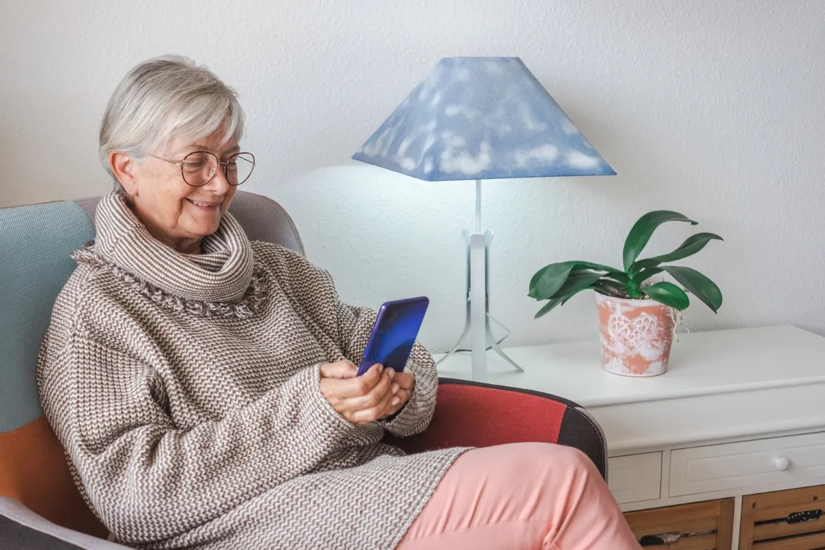 Ældre kvinde i strikket sweater sidder i en lænestol og smiler til sin mobiltelefon