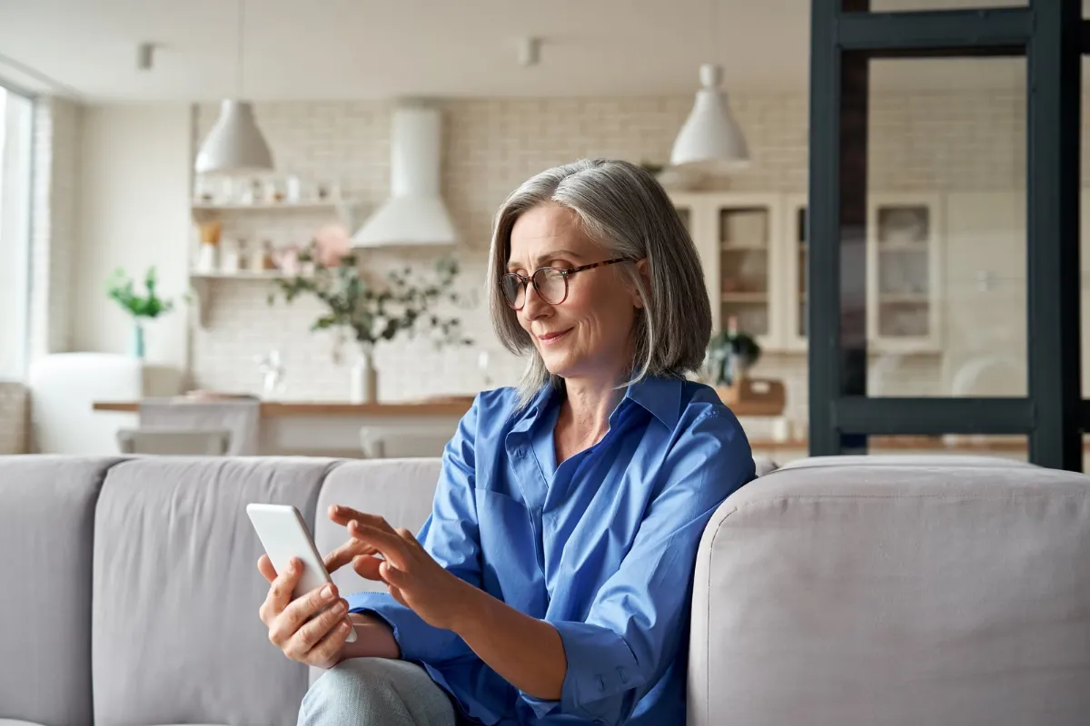 Kvinde med gråt hår og briller sidder i en lys sofa og kigger på sin mobiltelefon